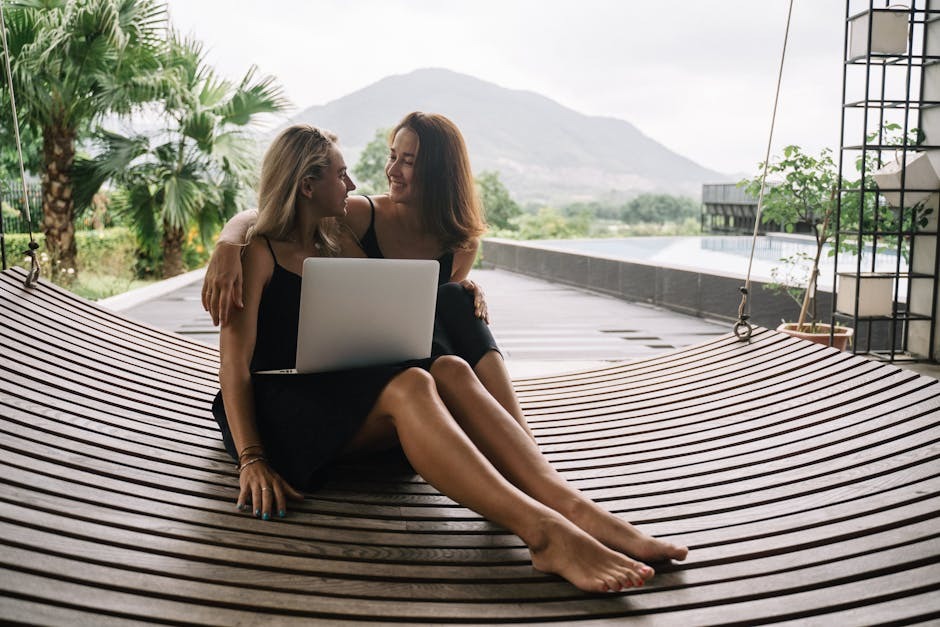 Two women relaxing outdoors reviewing dating profiles together on a laptop, illustrating joint verification by couples